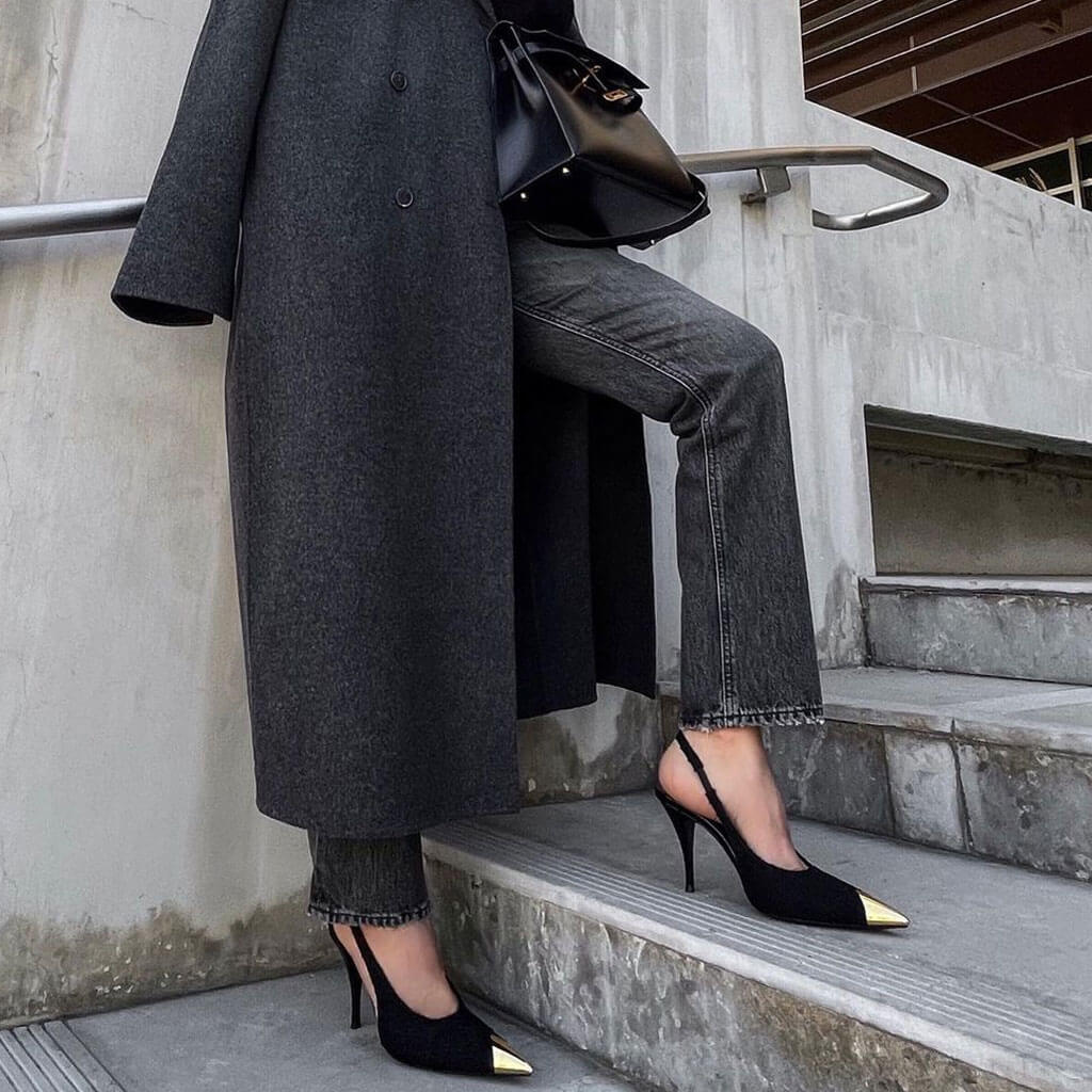 A person wearing black tweed slingback pumps with metallic toe caps and pointed toes. The person is seated on a staircase, with one foot placed on a step, demonstrating the heel height and style of the shoes.
