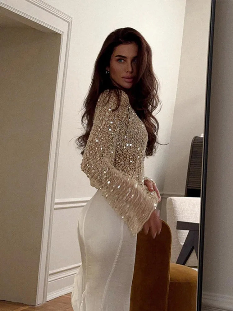 Woman wearing a sparkly top and white pants in a room with a mirror.