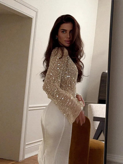 Woman wearing a sparkly top and white pants in a room with a mirror.