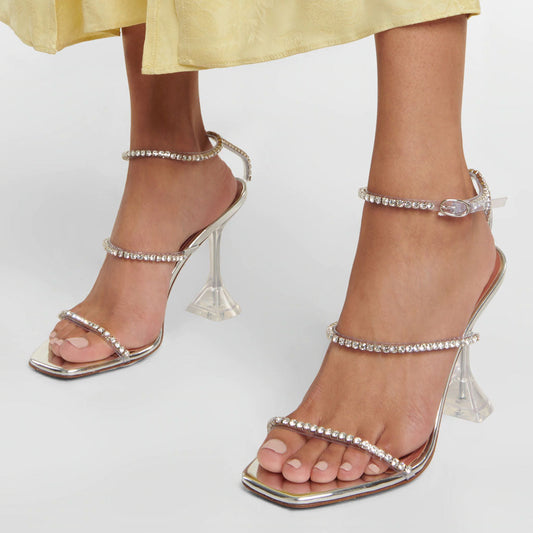 A pair of silver ankle strap sandals with square toes and clear PVC heels, adorned with shimmery rhinestone embellishments.